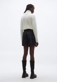 Bel pulover z rebrastim vzorcem in visokim ovratnikom, kombiniran s temnimi kratkimi jeans hlačami z zadnjimi žepi in vezenim logotipom, nošenimi s črnimi škornji do kolen.