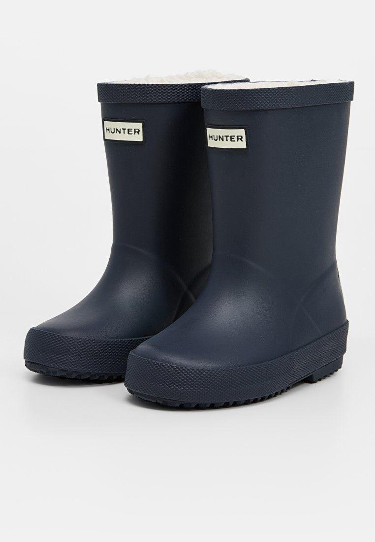 Marineblaue Gummiregenstiefel mit strukturierten Sohlen und weißem Fleece-Futter. Mit einem kontrastierenden Markenlabel an der Vorderseite zur Identifikation.