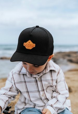 Fekete trucker sapka narancssárga gyémánt logóval, amelyet egy fehér kockás ingben lévő gyermek visel, ülve a homokban az óceán mellett.