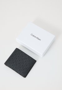 Portafoglio in pelle nera con un motivo testurizzato, con il logo Calvin Klein, posizionato accanto a una scatola bianca con il logo stesso sopra.