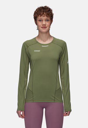 Personne portant un haut de sport à manches longues vert olive avec le logo "Mammut" et un pantalon ajusté mauve sur un fond uni.