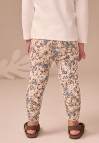 Leggings in velluto a coste con motivo floreale beige con accenti blu e marroni, abbinati a una maglietta bianca girocollo e sandali marroni.