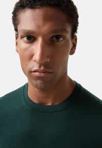 Ciemnozielony sweter typu crew-neck, dopasowany krój, gładka faktura, długie rękawy, minimalna ilość szwów, z miękkim, dzianinowym wykończeniem.