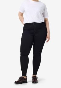 Personne portant un t-shirt blanc, un jean skinny noir taille haute et des chaussures à lacets marron foncé, debout avec une main dans le dos.