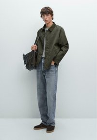 Groene katoen jack met een kraag, grijs t-shirt, lichtblauwe denim jeans en donkerbruine suède schoenen, met een bruine tas in de hand.