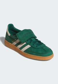 Green suede Adidas sneaker with white stripes, gum sole, laces, and a fold-over tongue covering the laces.