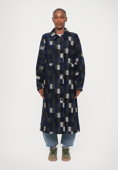 Manteau en laine à carreaux avec des carrés bleus, gris et noirs, doté d'un col, d'une fermeture à boutons et de poches avant ; associé à un pantalon en denim et des baskets.