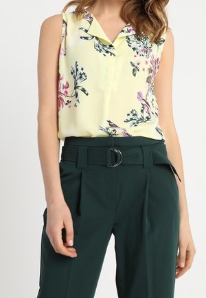 Chemisier sans manches jaune pâle avec des motifs floraux, assorti à un pantalon taille haute vert foncé avec une ceinture à boucle en argent.