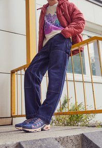 Pantalones cargo de mezclilla azul con bolsillos laterales, combinados con un suéter crema con un gráfico de montaña y una chaqueta acolchada roja. Zapatillas deportivas navy completan el conjunto.