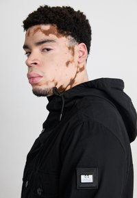 Sort hættetrøje med knap og snoretræk, brystlomme og mærke på ærmet. Modellen viser en sideprofil.