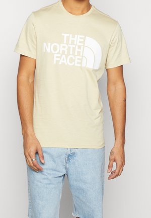 Mies, jolla on beige lyhythihainen T-paita, jossa on valkoinen "THE NORTH FACE" -logo, ja vaaleansiniset farkkushortsit.