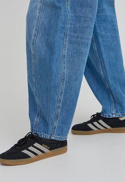 Zapatillas Adidas Gazelle de ante negro con suela de goma marrón, que presentan tres rayas blancas y la marca "GAZELLE" en dorado. Combinadas con unos pantalones de mezclilla azul claro.