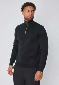 Pull noir zippé, tricot texturé, col haut, manches ajustées, et détail de fermeture éclair en argent. Porté avec un pantalon noir.
