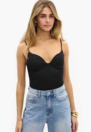 Frau mit langen blonden Haaren, die ein eng anliegendes schwarzes Spaghettiträger-Top und hellblaue hoch taillierte Jeans trägt, accessoriert mit goldenen Armbändern.