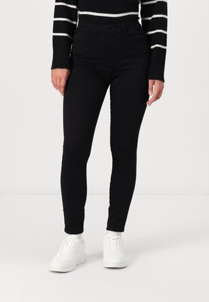 Femme portant un jean skinny noir, un pull noir à rayures horizontales blanches, des chaussettes blanches et des baskets blanches, debout sur un fond uni.