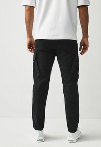 Schwarze Cargo-Hose mit zwei Seitentaschen, schmal zulaufendem Bein und dezenten Steppnähten, kombiniert mit einem locker sitzenden weißen T-Shirt und weißen Turnschuhen.