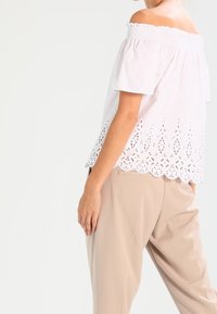 Vit off-shoulder topp med korta ärmar, prydd med en smockad halsringning och en intrikat hålmönster nederkant. Kombinerad med beige byxor.