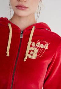 Punainen samettinen vetoketjullinen huppari, jossa on vaaleanbeiget nyörit, ja jossa on vaaleanbeige brodeerattu teksti ja numerot rinnassa, sileä pinta.