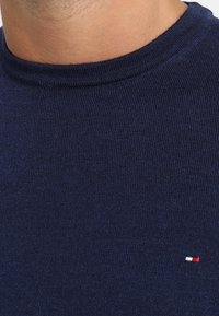 Mörkblå stickad tröja med rund halsringning och en liten logotyp i röd, vit och blå på bröstet. Mjuk textur, lättviktigt tyg.