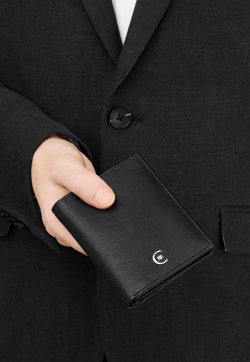 Mano con chaqueta de traje negra sosteniendo una cartera de cuero negro con textura, cerrada, con un pequeño logotipo plateado en la esquina.