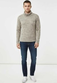 Suéter de cuello alto texturizado de color gris claro con un ajuste relajado, combinado con jeans azul oscuro y zapatillas blancas, posando frente a un fondo neutro.