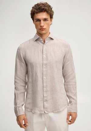 Młody mężczyzna z kręconymi włosami, ubrany w jasnobeżową lnianą koszulę na guziki z długim rękawem oraz białe spodnie na jednolitym tle.