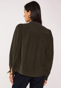 Blusa in velluto a coste verde scuro con maniche a sbuffo, pieghe posteriori e polsini abbottonati. Presenta un tessuto texturizzato e un design classico del collo.