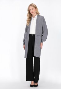 Grauer langer Cardigan aus weichem Stoff mit zwei Fronttaschen, offenem Design und gerippten Bündchen, kombiniert mit schwarzen weiten Hosen.
