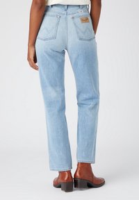 Persona vistiendo jeans rectos Wrangler de color azul claro y botas de cuero marrón hasta el tobillo, de pie contra un fondo sencillo.