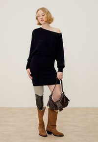 Kobieta z krótkimi blond włosami, ubrana w czarną sukienkę z odkrytymi ramionami, brązowe kowbojki, trzyma brązową torebkę i ma protezę lewej nogi.