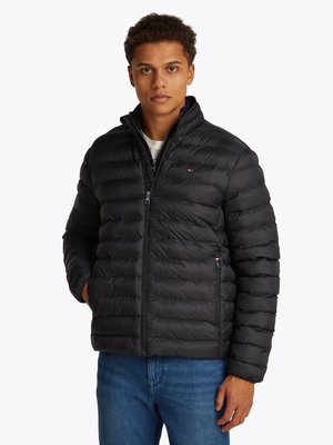 Casaco puffer preto com textura acolchoada e gola alta. Apresenta uma frente com fecho éclair e bolsos laterais. Usado por cima de uma camisa branca e jeans azuis.