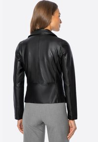 Chaqueta de cuero negro con un diseño ajustado, que presenta un cuello con muescas y costuras en los hombros. Llevada sobre pantalones grises, mostrando una silueta elegante.