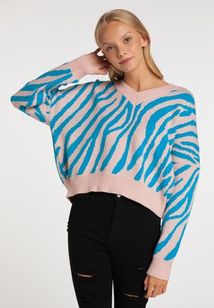 Junge Frau trägt einen rosa-blau gestreiften Pullover mit V-Ausschnitt und schwarze zerrissene Jeans, posiert mit einer Hand im Haar.