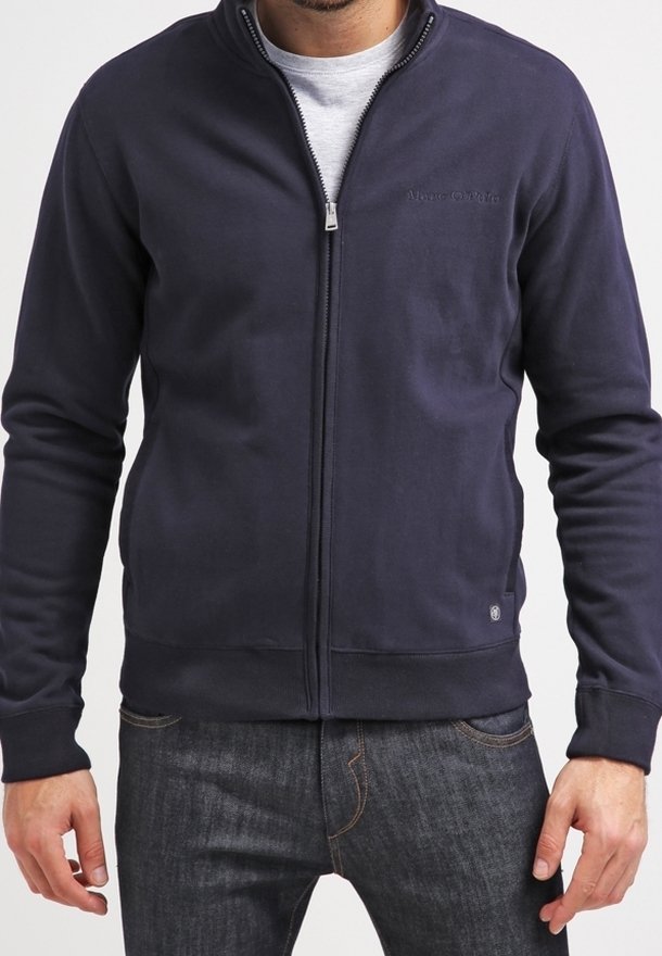 Homme portant un sweat-shirt bleu marine à fermeture éclair sur un t-shirt gris clair et un jean bleu foncé, debout les bras détendus le long du corps.