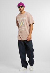 Tricou din bumbac roz cu grafică „Money Talks” și croială supradimensionată, asortat cu blugi donker bleumarin și adidași albi, purtat cu o caciulă mov.