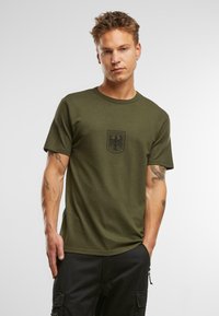 Olivza zaļa kokvilnas T-kreklā ar īsām piedurknēm un apaļu kakla izgriezumu. Uz krūtīm ir melns izšūts ērgļa emblēma. Gluda tekstūra.