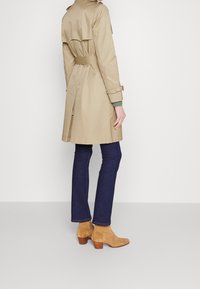 Beżowy trencz z paskiem w talii, długimi rękawami i rozcięciem z tyłu, zestawiony z ciemnymi jeansami i beżowymi botkami za kostkę. Gładka faktura materiału.