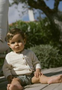 Bijela pletena cardigan jakna s drvenim gumbima, uparena sivoj šortse. Dijete sjedi na drvenoj površini, ima kovrčavu kosu i bose noge.