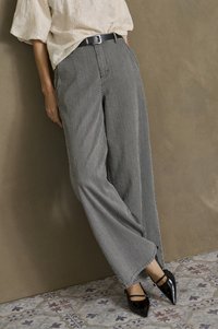 Henkilö, jolla on ylhäältä korkeat, raidalliset leveälahkeiset housut mustalla vyöllä, vaalea teksturoitu paita ja mustat teräväkärkiset Mary Jane -kengät, seisoo kuviollisella lattialla.