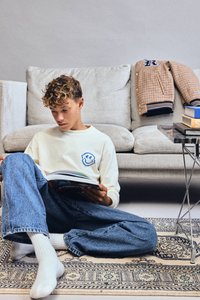 Nastolatek z kręconymi włosami siedzi na wzorzystym dywanie, czytając książkę, ubrany w białą bluzę i niebieskie dżinsy, z beżową kanapą i kurtką w tle.