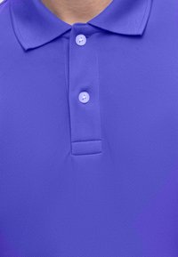 Gros plan d'une personne portant un polo violet avec un col replié et deux boutons blancs attachés au niveau de l'encolure.