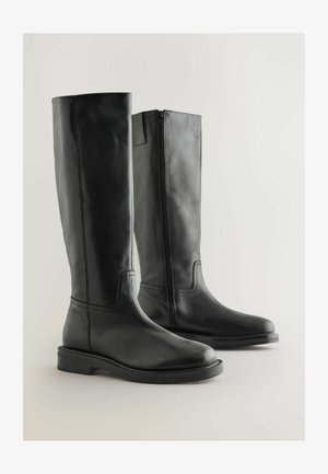 Bottes noires montantes en cuir lisse. Présentent un bout arrondi, des coutures minimales, une semelle plate, et une fermeture éclair sur le côté pour un enfilage facile.
