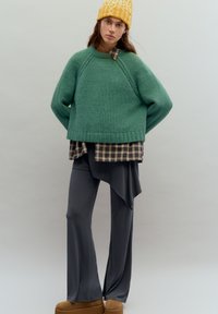 Ylisuuri vihreä neulepaita, kerrostettuna ruskean ruudullisen paidan päälle, yhdistetty tummaan harmaaseen leveälahkeiseen housuun ja vaaleanruskeisiin korollisiin saappaisiin. Keltainen teksturoitu pipo.