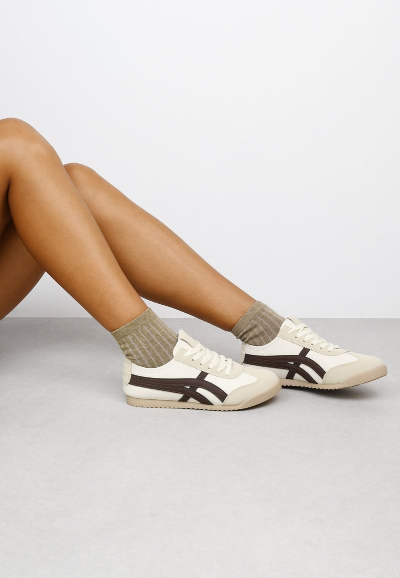 Elegante Sportschuhe in Creme und Braun, mit einem glatten Obermaterial, kontrastierenden Streifen und einer strukturierten, beigefarbenen Sohle. Kombiniert mit gestreiften olivfarbenen Socken.