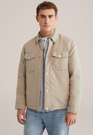 Beige padded jacket with snap buttons and two chest pockets. Worn over a light gray shirt, featuring a smooth texture and casual design.