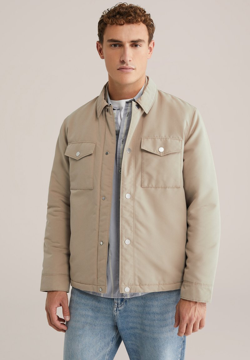 Beige gewatteerde jas met drukknoopsluitingen en twee borstzakken. Draag over een lichtgrijs shirt met een gladde textuur en een casual ontwerp.