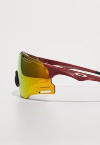 Czerwone sportowe okulary przeciwsłoneczne z gradientowymi żółtymi soczewkami, wyróżniające się unikalnym kanciastym wzornictwem, lekką ramką i srebrnym akcentem na skroni.