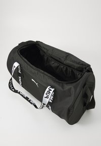 Sac de sport noir avec un extérieur en tissu lisse, deux poignées supérieures et une fermeture éclair. Il possède un logo et des sangles à motifs. L'intérieur est spacieux.