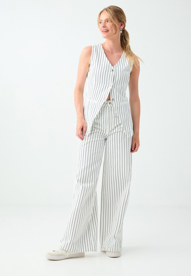 Femme aux cheveux blonds attachés en queue de cheval, portant un gilet blanc sans manches à fines rayures et un pantalon large assorti avec des baskets blanches.