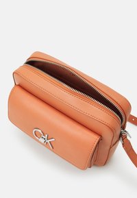 Oranžová kožená kabelka přes rameno s uzávěrem na zip, předním klopou, stříbrnými kovovými doplňky a vyraženým logem. Kompaktní a pevná konstrukce.